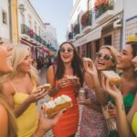 A group of women enjoying hen party activities in the sunny cobblestone streets of Albufeira Old Town, surrounded by white buildings and colourful flowers.