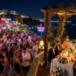 A night scene showing a lively outdoor bar with people socialising and dancing next to a quiet, candlelit private dinner gathering by the coast.